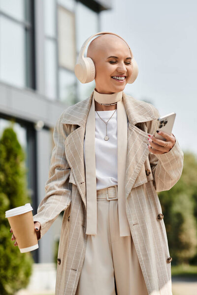 A young bald woman in stylish clothing smiles joyfully, holding a coffee cup and a phone.