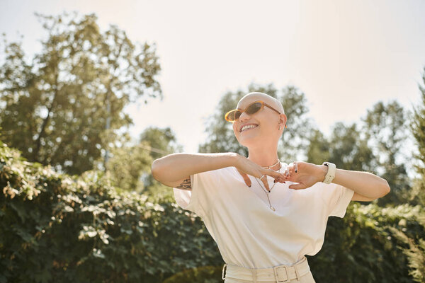 A joyful young bald woman dances in elegant clothing under the warm sun amid lush greenery.