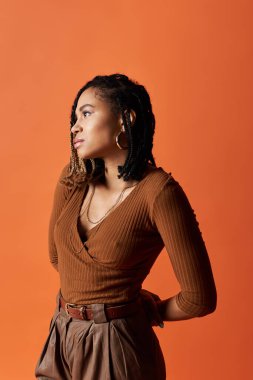 Saçları örgülü genç bir kadın stilini yaratıcı bir stüdyoda parlak turuncu bir arka planda sergiliyor..