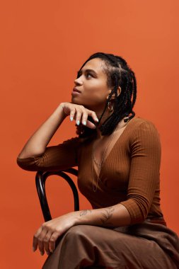 Örgülü saçları olan genç bir kadın parlak turuncu bir arka plana karşı düşünceli bir şekilde poz verir..