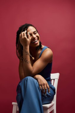 Saçları örgülü genç, modaya uygun Afrikalı Amerikalı bir kadın renkli bir stüdyo arkaplanında neşeli bir şekilde gülümsüyor..