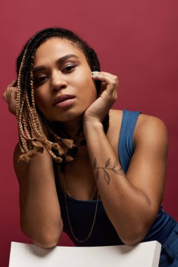 Örgülü saçları olan kendine güvenen bir kadın renkli bir stüdyoda poz veriyor, şık kıyafetlerini ve tavırlarını sergiliyor..