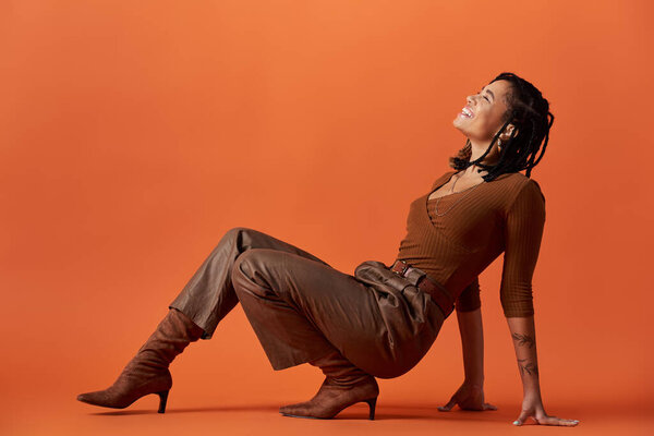 Молодая женщина с плетеными волосами сидит изящно, излучая уверенность и стиль на ярком студийном фоне.