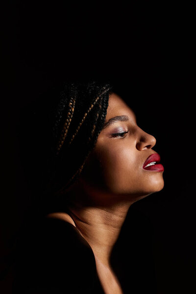 A young African American woman with vibrant red lips styles braided hair, showcasing her fashion sense in a studio.