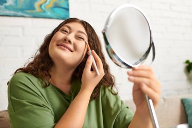 Kıvırcık saçlı genç bir kadın, oturma odasında makyaj yaparken gülümsüyor..