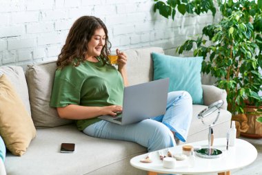 Neşeli genç bir kadın, şık bir yaşam alanında dizüstü bilgisayarında çay içerken güzellik rutinini keşfediyor..