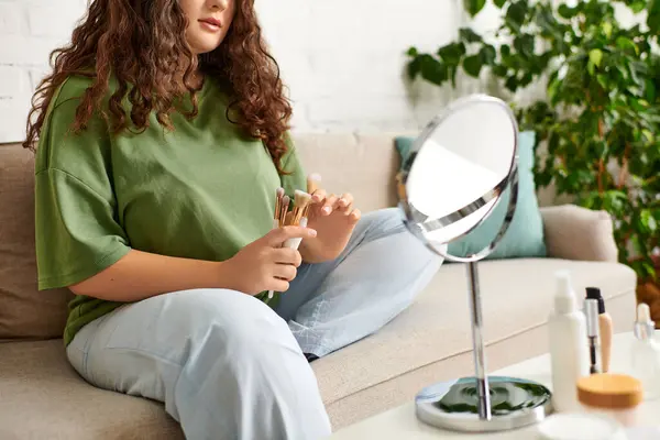 stock image A young woman with curly hair explores her beauty products while relaxing on a cozy living room sofa.