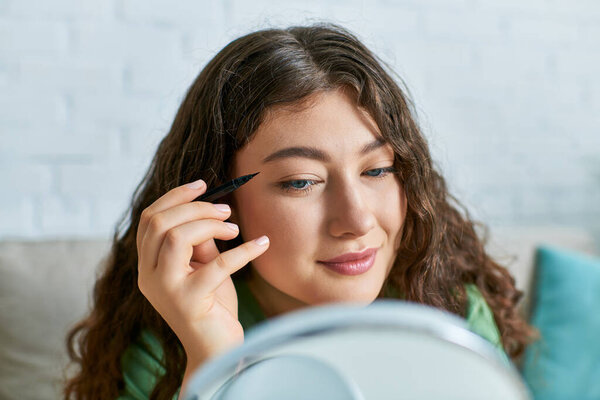A cheerful young woman with curly hair applies cosmetics, embracing her beauty routine in a cozy space.