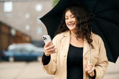 Siyah elbiseli ve bej renkli trençkotlu genç bir kadın telefonuna bakarken gülümsüyor..