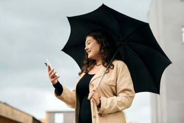 Güzel genç bir kadın, sonbaharda siyah bir şemsiyenin altında gülümseyip telefonunu kontrol ediyor..