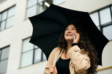 Sonbahar yağmuru sırasında şemsiyesinin altında telefonunda kıvırcık saçlı neşeli, iri yarı bir kadın sohbet ediyor..