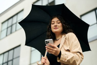 Havalı siyah elbiseli ve trençkotlu iri yarı bir kadın dışarıda sonbaharın tadını çıkarırken telefonunu kontrol ediyor..