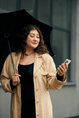 Genç, iri yarı bir kadın, şık siyah bir kıyafet ve rahat bir trençkot giymiş, telefonuna gülümsüyor..