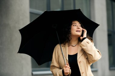 Şık, iri yarı bir kadın dışarıda yürüyor, telefonda şemsiyeyle kendini koruyor..