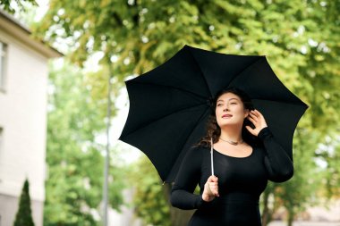 Siyah elbiseli neşeli, iri yarı bir kadın şemsiyesinin altında sonbahar atmosferini kucaklayarak dışarıda yürüyor..
