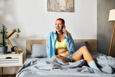 Neşeli genç bir kadın, rahat yatağında gündelik kıyafetleriyle telefonla eğleniyor..
