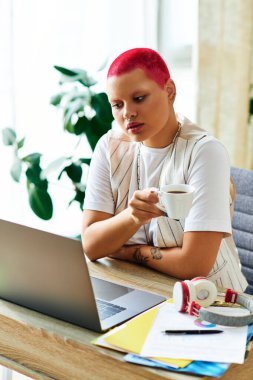 A young woman with vibrant hair sips coffee, focused on her laptop in a cozy workspace. clipart