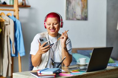 A vibrant young woman smiles as she balances her phone and a pencil, immersed in her creative space. clipart