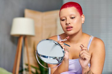 In a cozy living space, a young woman focuses on her reflection as she carefully applies lipstick. clipart