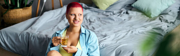 A young woman with short red hair sits comfortably at home, sipping a drink joyfully.