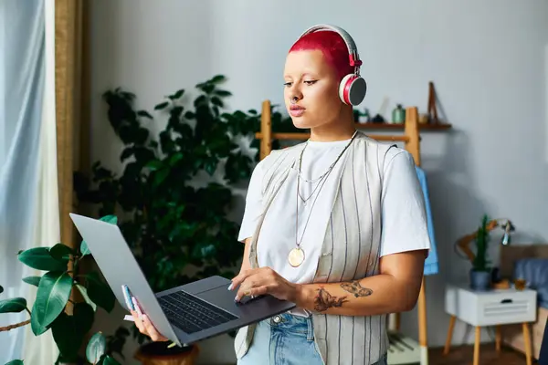 stock image A young woman focused on her laptop at home, wearing headphones and surrounded by greenery.