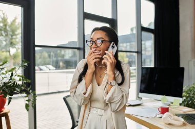 A young woman in glasses beams while talking on her phone in a bright office setting. clipart