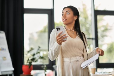 Profesyonel kıyafetli genç bir kadın telefonunu ve belgelerini düşünceli bir şekilde bir ofiste tutuyor..