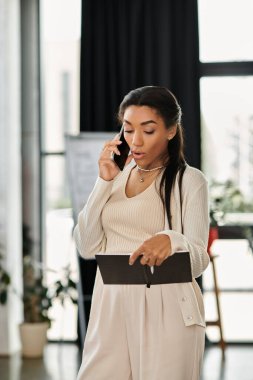 Genç bir kadın ofis ortamında notlarını kontrol ederken telefon görüşmesine odaklanır..