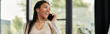 A cheerful young woman engages in a phone conversation, radiating positivity in her workspace. clipart