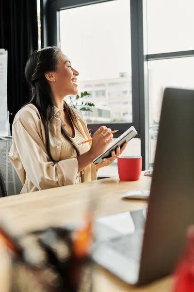 stock image Inside a bright office, a young woman joyfully takes notes while enjoying a warm beverage.