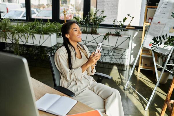 Молодая женщина улыбается, сидя в своем современном офисе, используя свой телефон и наслаждаясь моментом.