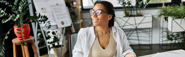 A cheerful young woman works attentively in a stylish office filled with plants and light.