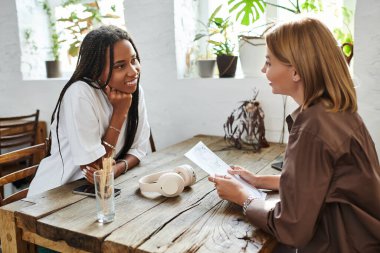 Two women share smiles and laughter over drinks at a charming cafe filled with greenery. clipart