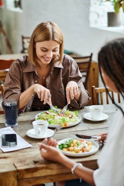 İki kadın, hareketli bir kafede keyifli bir yemeği paylaşıyor, gülüyorlar ve birbirlerine eşlik ediyorlar..