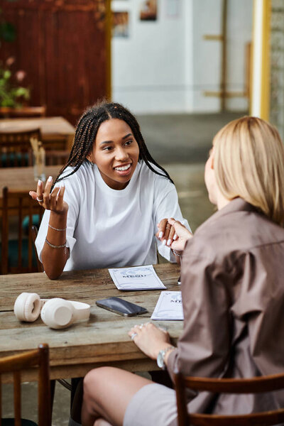 A cheerful African American woman with braided hair smiles warmly at her girlfriend during a cafe date.