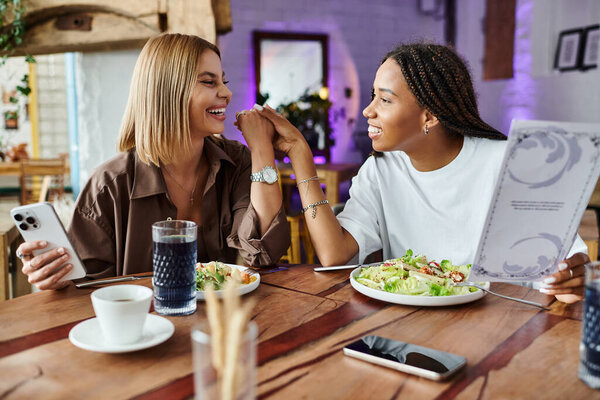 Радостная мультикультурная пара делится светлым моментом в кафе, наслаждаясь вкусной едой и компанией друг друга.