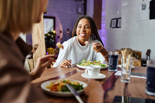 В очаровательном кафе, радостная афроамериканка делится смехом и едой со своей девушкой, создавая воспоминания.