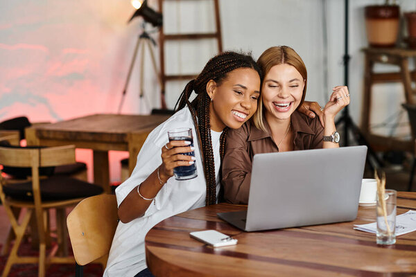 Счастливая многонациональная пара делится смехом и напитками в кафе, занимаясь совместным ноутбуком.