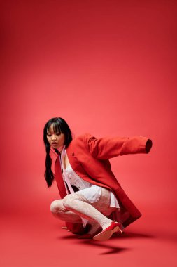 A poised Asian woman crouches stylishly, showcasing bold fashion choices in a vibrant setting.