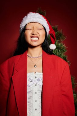 A cheerful Asian woman is celebrating the holiday spirit in a striking red jacket and Santa hat. clipart