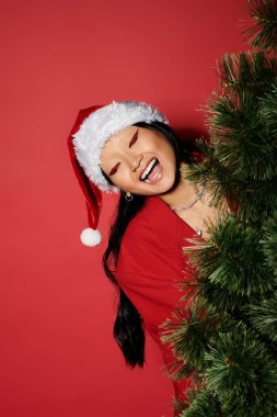 Kırmızılı neşeli bir kadın ve Noel Baba şapkası Noel ağacının arkasından neşeli bir şekilde dikizliyor..