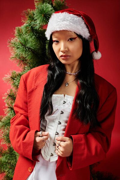 Элегантная азиатка в красном пальто и шляпе Санты воплощает дух праздника рядом с елкой.