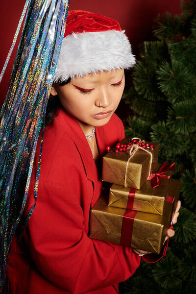 Эта веселая женщина, украшенная шляпой Санты, обнимает элегантные подарки среди праздничных украшений.