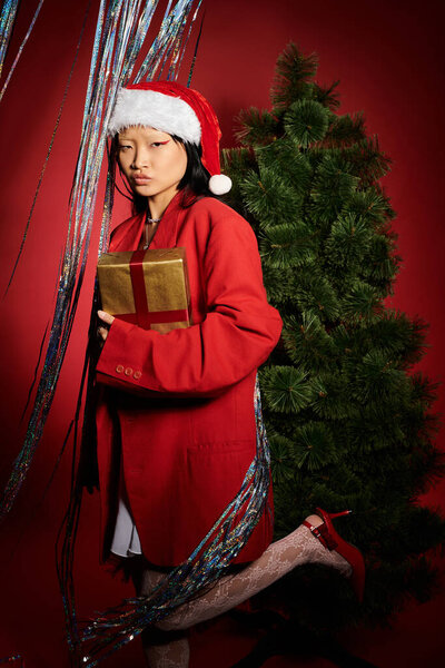 Элегантная женщина позирует с подарком, окруженный праздничными украшениями и фоном рождественской елки.