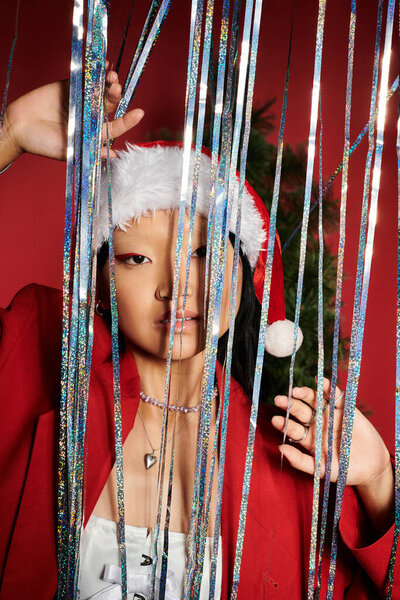 A captivating Asian woman radiates holiday spirit, peeking through colorful decorations.