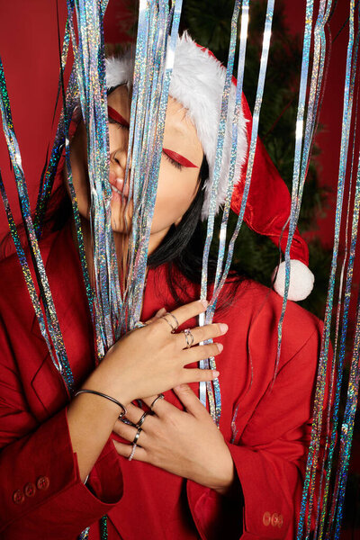 Элегантная женщина в шляпе Санты игриво позирует среди сверкающих праздничных лент, излучающих радость.