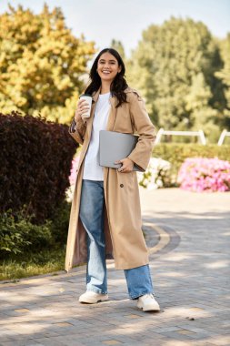 A cheerful young woman strolls through a vibrant autumn park, holding a coffee and laptop. clipart