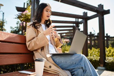 Dressed warmly for autumn, a young woman sits on a bench, engaged in her work outdoors. clipart