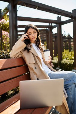 A stylish young woman sits on a park bench, sipping coffee while focused on her laptop. clipart