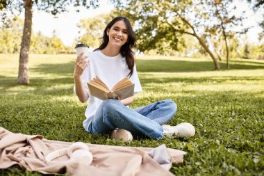 Kalın giysiler giyen bir kadın, sonbahar güneşinde parkta rahatlar, kitap okur ve kahvesini yudumlar..